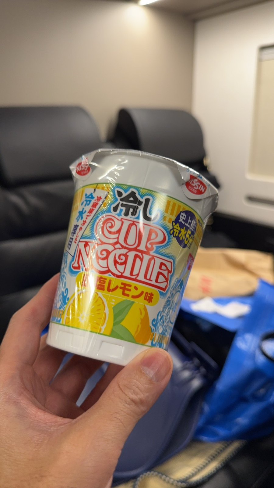 ソファの置かれた室内で、日清食品の「冷しカップヌードル 塩レモン味」の未開封カップを手に持っているところ。ふたには「冷水専用」「史上初 冷水5分」と書かれている。