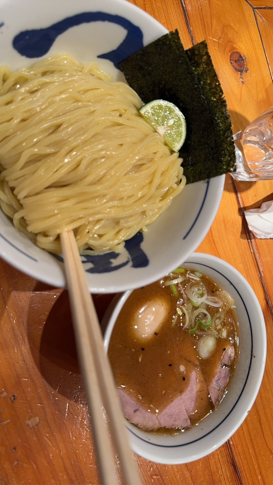 青い絵付けの丼に盛られた太めのつけ麺。脇に海苔2枚とすだちの輪切りが添えられ、手前の小丼には味玉・チャーシュー・刻み葱が入った茶色いつけ汁が置かれている。木のテーブルに箸が渡してある。